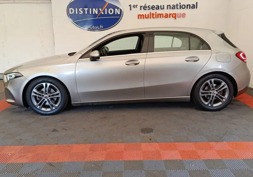 Vue de profil côté gauche d'une Mercedes Classe A 180 Business Line gris clair, avec jantes alliage et feux arrière LED allumés.