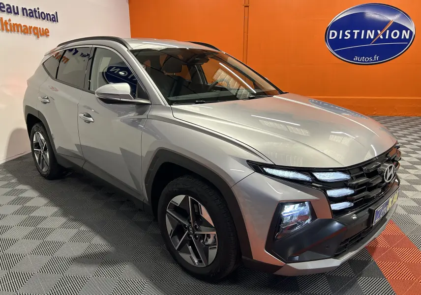 Hyundai Tucson 2025 hybride en Shimmering Silver, vue 3/4 avant droit avec jantes 18 pouces et phares LED allumés.