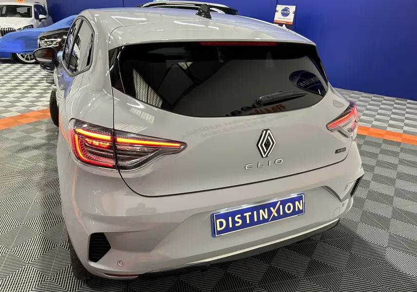 Vue 3/4 arrière droite d'une Renault Clio V E-Tech gris Rafale métal avec feux arrière allumés en intérieur showroom.