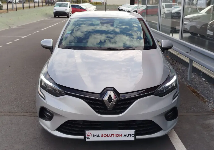 Vue avant d'une Renault Clio V gris platine, avec calandre noire et phares LED allumés, stationnée devant un bâtiment vitré.