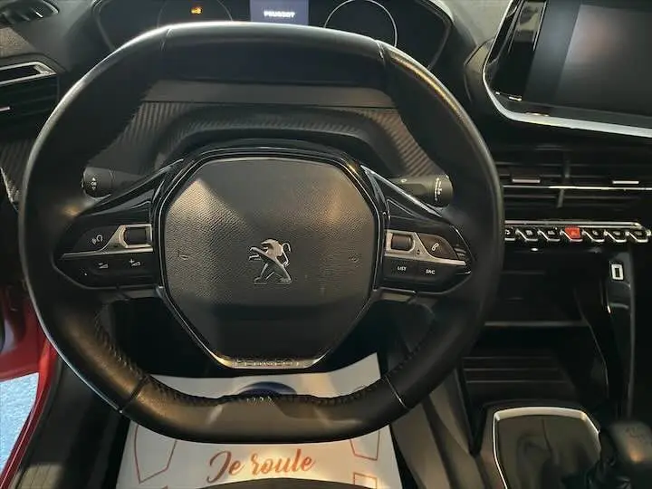 Vue rapprochée du volant multifonction noir avec logo Peugeot, tableau de bord et partie de la console centrale du 208 rouge Elixir.