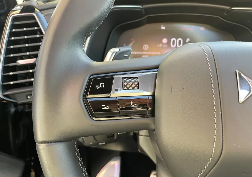 Gros plan sur la partie gauche du volant en cuir noir du DS7 2024, avec commandes intégrées et tableau de bord numérique flou en arrière-plan.