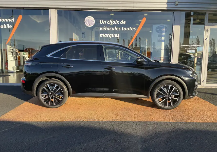 Profil côté gauche d'un DS7 noir Perla Nera 2024 avec jantes bi-ton et vitres teintées, devant une vitrine commerciale.
