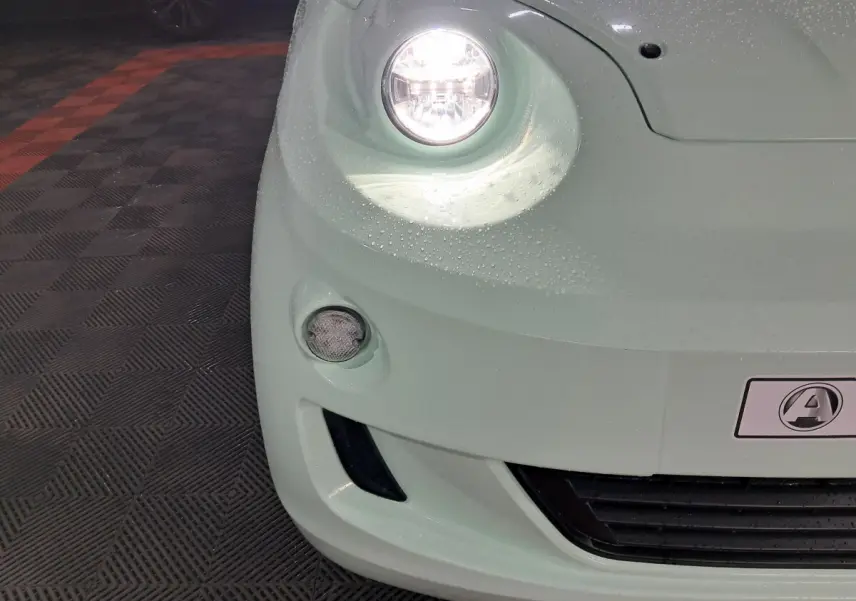 Gros plan sur l'avant gauche vert d'une Aixam MINAUTO E chic 2025 avec phare allumé et logo visible.