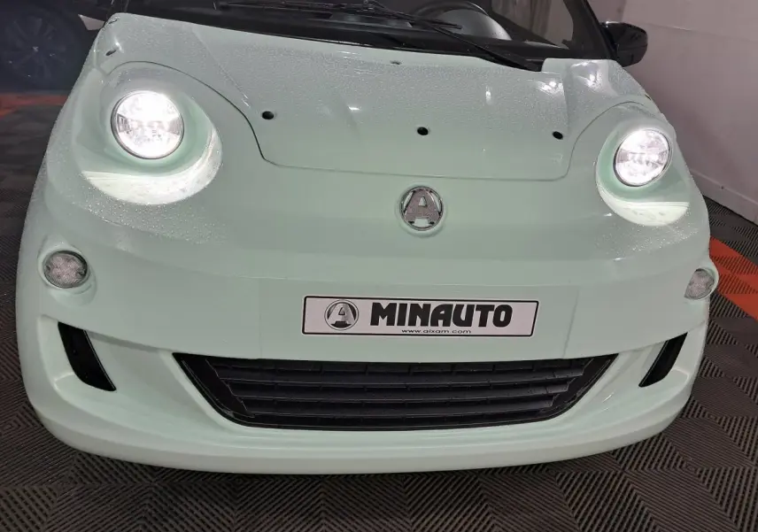 Vue de face d'une Aixam MINAUTO E chic 2025 vert clair avec phares allumés et logo centré sur le capot.