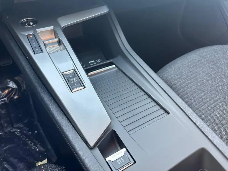 Vue rapprochée de la console centrale grise de la Peugeot 308 2025 avec boîte auto et bouton Drive Mode, siège tissu gris visible.