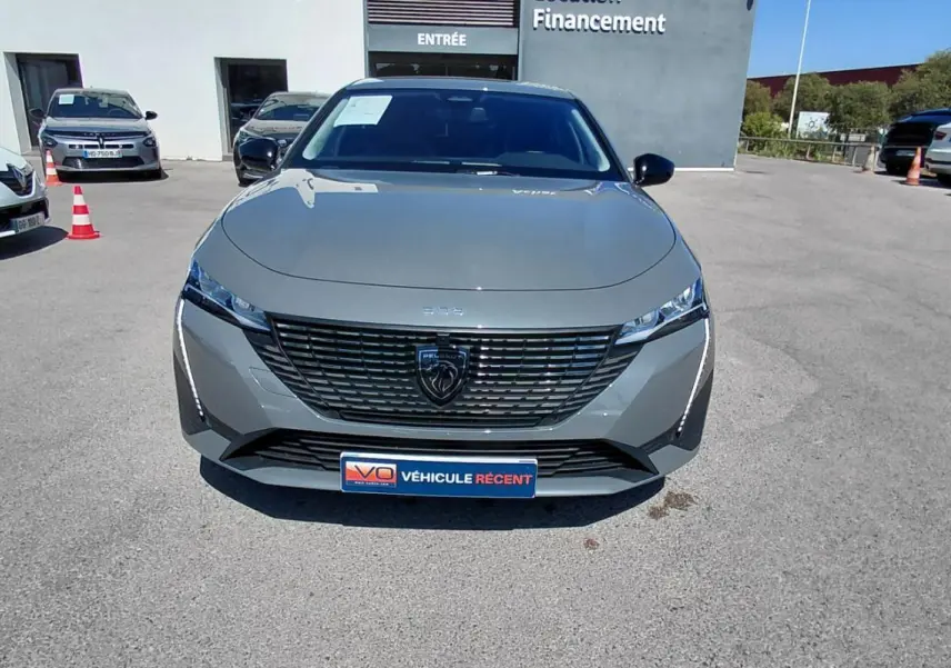 Vue avant d'une Peugeot 308 Hybrid 145 gris métallisé avec calandre noire et feux LED fins, stationnée en extérieur.