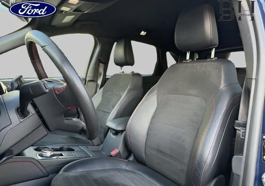 Vue intérieure côté conducteur du Ford Kuga 2023 bleu, sièges sport noirs avec surpiqûres rouges et volant cuir perforé.