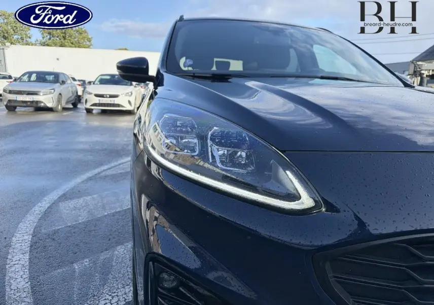 Gros plan sur l'avant gauche du Ford Kuga bleu avec phares LED et calandre noire brillante sous la pluie.