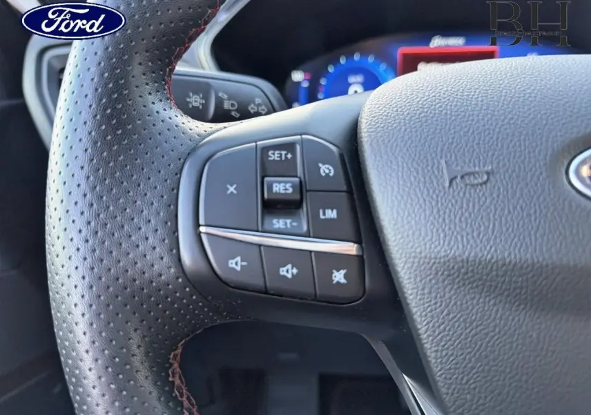 Gros plan sur le volant cuir perforé du Ford Kuga 2023 avec commandes multifonctions côté gauche et tableau de bord numérique bleu.