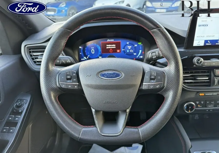 Vue centrée sur le volant cuir perforé avec surpiqûres rouges du Ford Kuga bleu, tableau de bord numérique et écran tactile visible.