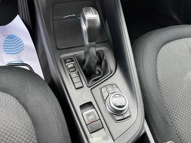Vue intérieure rapprochée sur la console centrale noire du BMW X1 20D, montrant levier de vitesse et commandes multimédia.