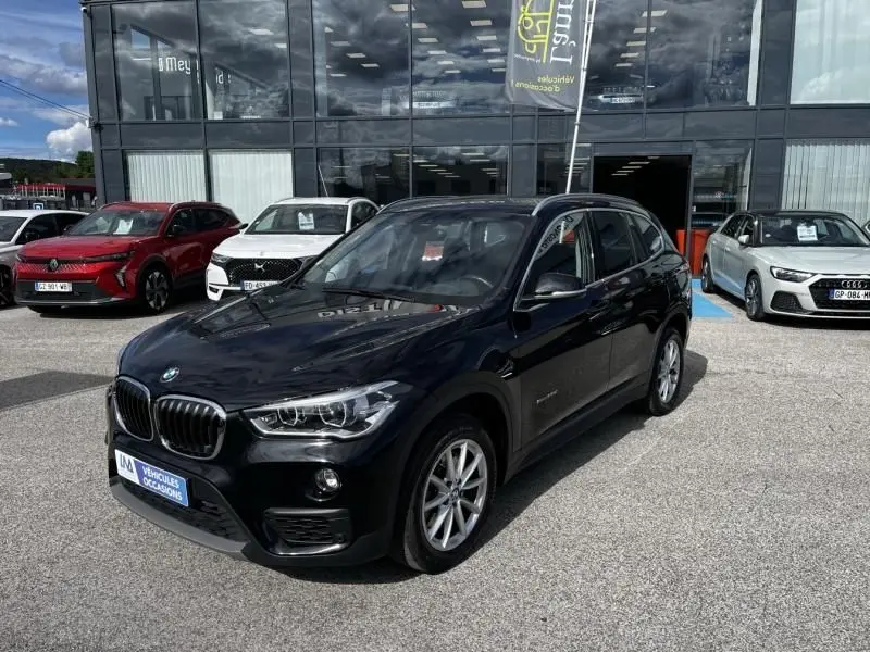 BMW X1 20D noir métal en 3/4 avant droit, avec calandre caractéristique et jantes alliage visibles, stationné devant un showroom.