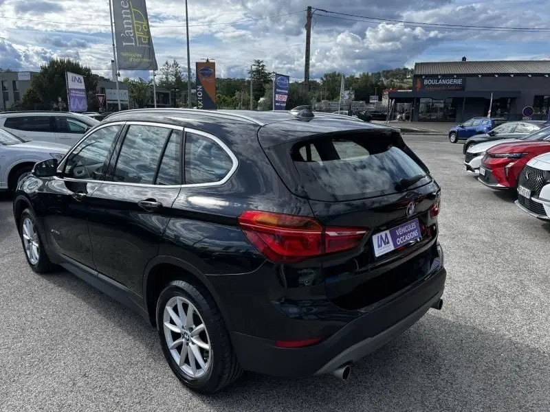 BMW X1 20D noir métal vue 3/4 arrière droit sur parking avec jantes alliage et feux arrière allongés.