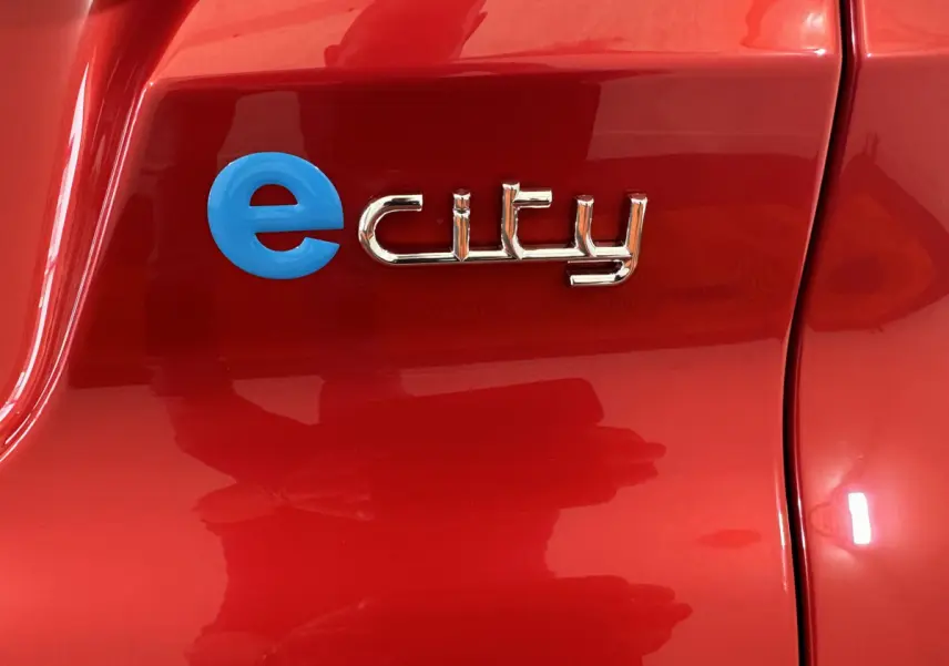 Gros plan sur l’inscription e city en bleu et chrome sur la carrosserie rouge de l’AIXAM AMBITION E CITY PACK 2025 électrique.