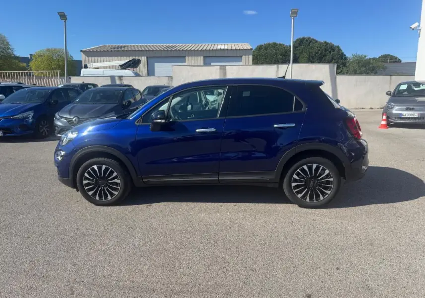Vue de profil côté gauche d'un FIAT 500X bleu 2024 avec jantes classiques noires et chromées sur parking extérieur.