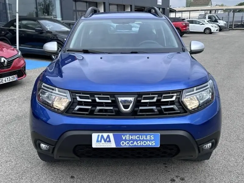 Vue de face d'un Dacia Duster 2022 bleu métal, avec calandre noire et phares allumés, sur parking extérieur.