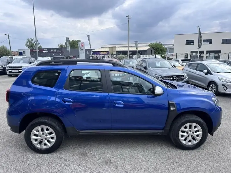 Vue de profil côté gauche d'un Dacia Duster bleu métal 2022 avec barres de toit et jantes argentées.