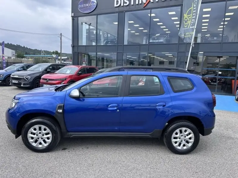 Vue de profil côté gauche d'un Dacia Duster bleu métal 2022 stationné devant un showroom, avec jantes argentées et rétroviseurs gris.