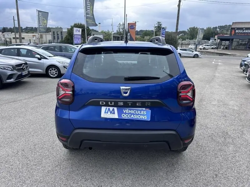 Vue arrière d'un Dacia Duster bleu métal 2022 avec logo et plaque "Véhicules Occasions" sur un parking extérieur.