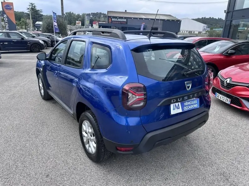 Vue 3/4 arrière droite d'un Dacia Duster bleu métal 2022, version ECO-G 1.0 Confort 4x2, avec barres de toit visibles.