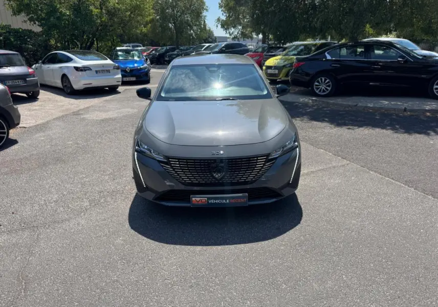Vue avant frontale d'une Peugeot 308 gris métallisé 2025 avec calandre noire et logo central sur un parking.