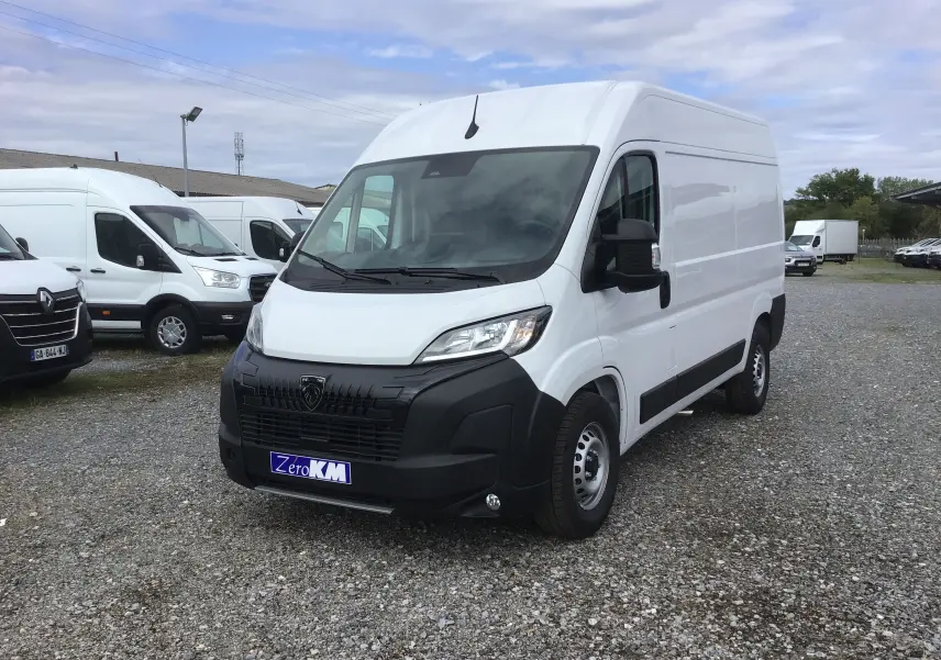 Fourgon Peugeot Boxer blanc en 3/4 avant droit sur parking gravillonné avec plusieurs véhicules utilitaires en arrière-plan.