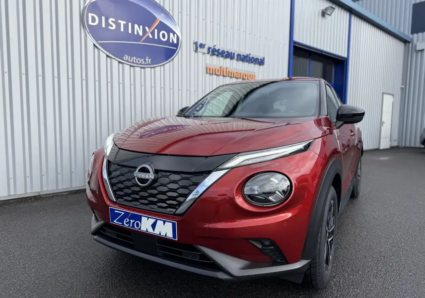 Nissan Juke 1.6 Hybrid rouge Fuji en 3/4 avant droit, avec calandre noire et phares ronds distinctifs.