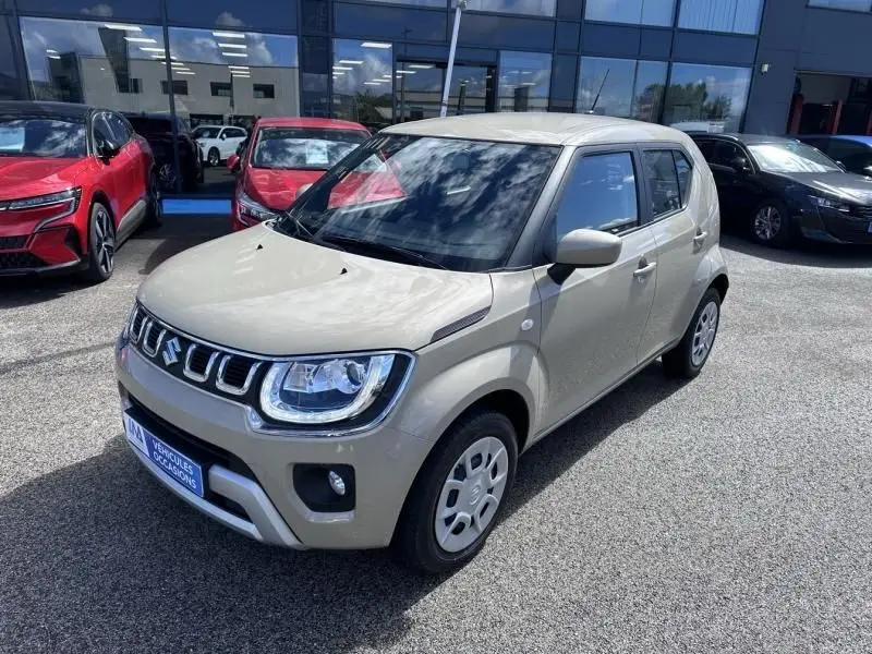 Suzuki Ignis beige vue 3/4 avant droit, avec calandre chromée et phares LED, stationnée devant un bâtiment vitré.