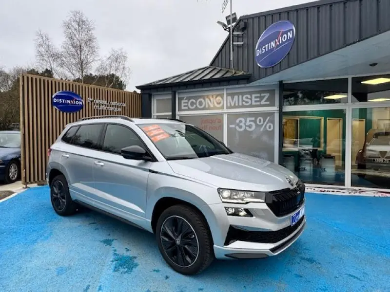 Skoda Karoq gris diamant métallisé en 3/4 avant droit, avec toit panoramique et jantes noires, devant un concessionnaire.