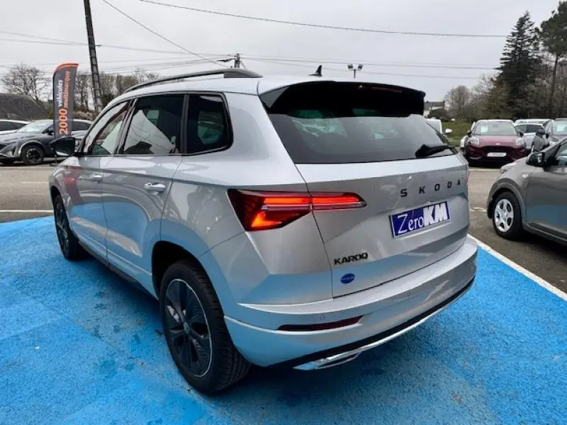 Vue 3/4 arrière droite d'un Skoda Karoq gris diamant métallisé avec feux arrière LED allumés sur parking.