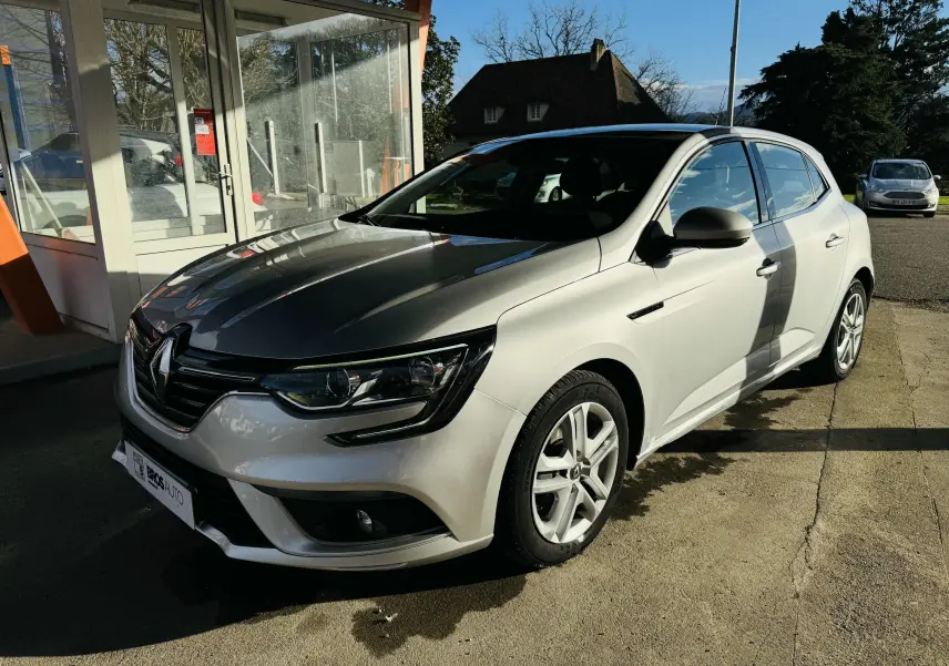 Renault Mégane IV berline gris vue 3/4 avant droit, stationnée devant un bâtiment vitré par temps ensoleillé.