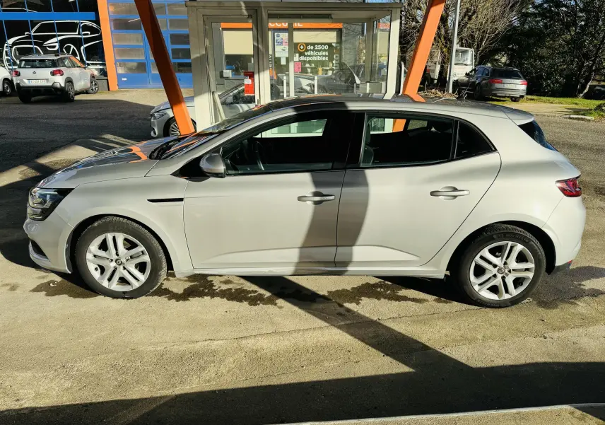 Profil côté gauche d'une Renault Mégane IV Berline gris clair garée en extérieur sous un auvent orange.