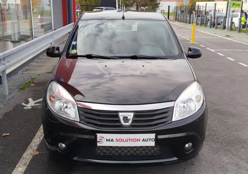 Vue frontale d'une Dacia Sandero noire de 2011 avec calandre chromée et phares allumés sur un parking urbain.