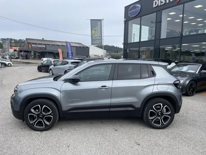 Vue de profil côté gauche d’un Jeep Avenger gris métal 2025 avec jantes noires et toit noir, stationné devant un bâtiment commercial.