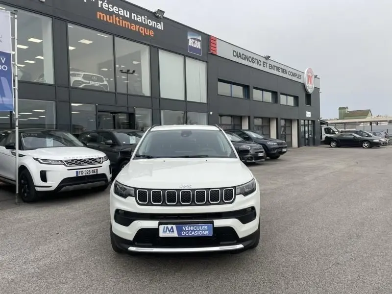 Vue avant d'un Jeep Compass blanc 2023, avec calandre distinctive à sept fentes, sur un parking devant un garage multimarque.
