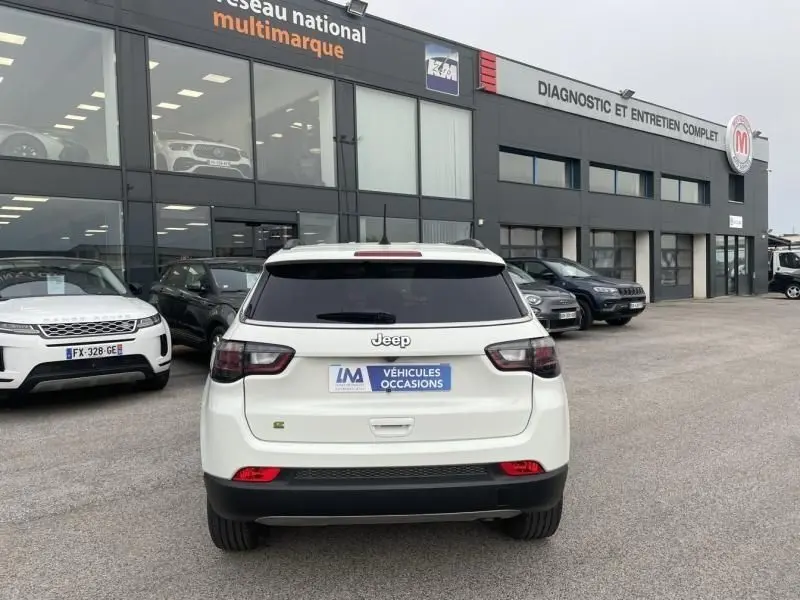 Vue arrière d'un Jeep Compass blanc 2023 avec badge Limited et logo Jeep, stationné devant un garage multimarque.