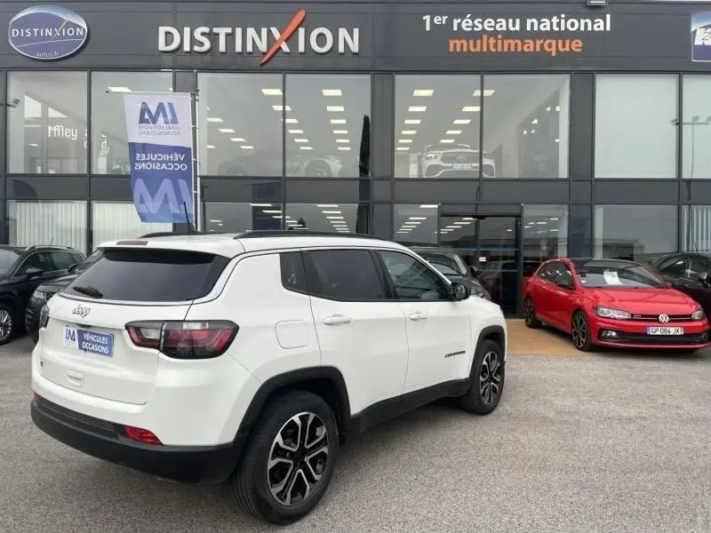 Vue 3/4 arrière droite d’un Jeep Compass blanc 2023 avec jantes noires et toit noir devant un showroom multimarque.