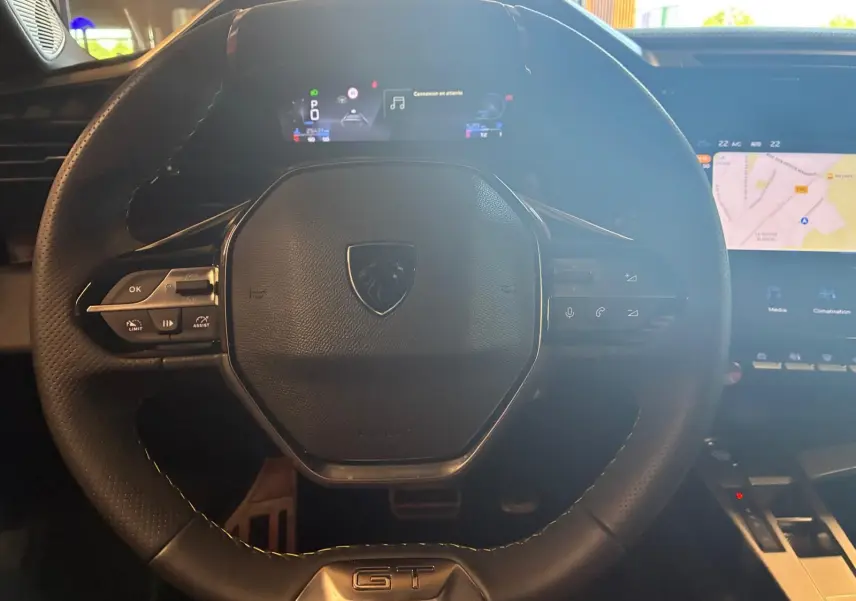 Vue rapprochée du volant cuir noir avec logo Peugeot et badge GT, tableau de bord numérique et écran tactile de navigation visibles.