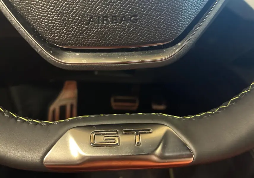 Gros plan sur le volant noir avec surpiqûres vertes et logo GT de la Peugeot 308 1.2 Hybrid 136 E-DCS6 GT.