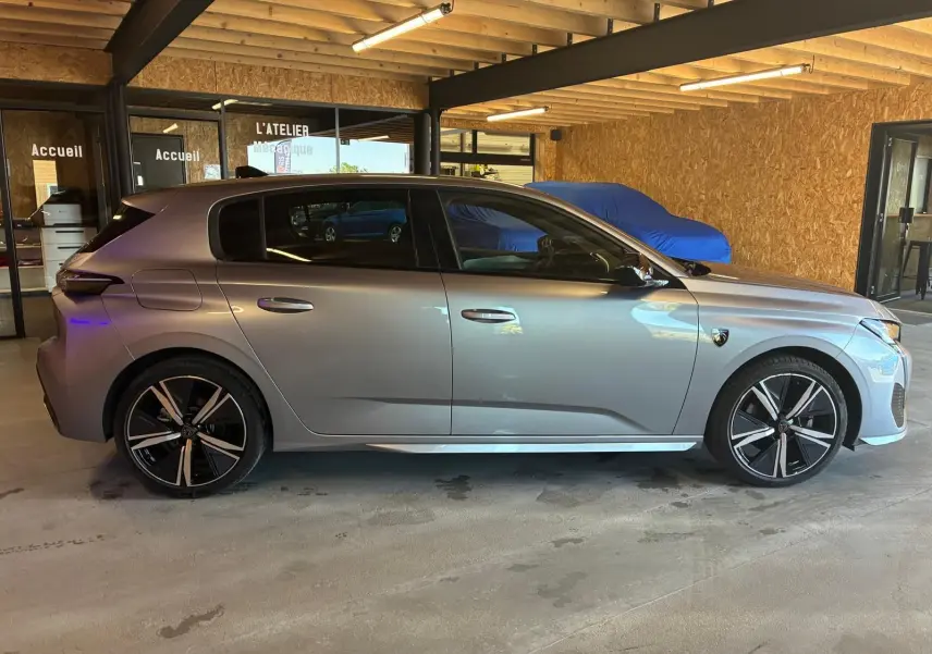 Profil droit d'une Peugeot 308 1.2 Hybrid 136 gris, avec jantes noires et détails sportifs, stationnée en intérieur.