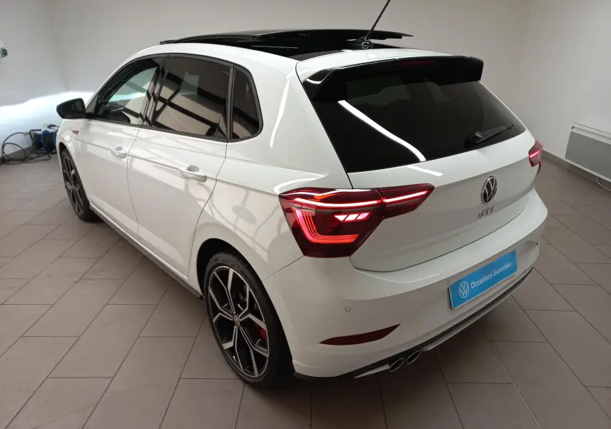 Vue 3/4 arrière droite d'une Volkswagen Polo GTI 2024 blanche avec toit noir ouvrant et feux arrière allumés.