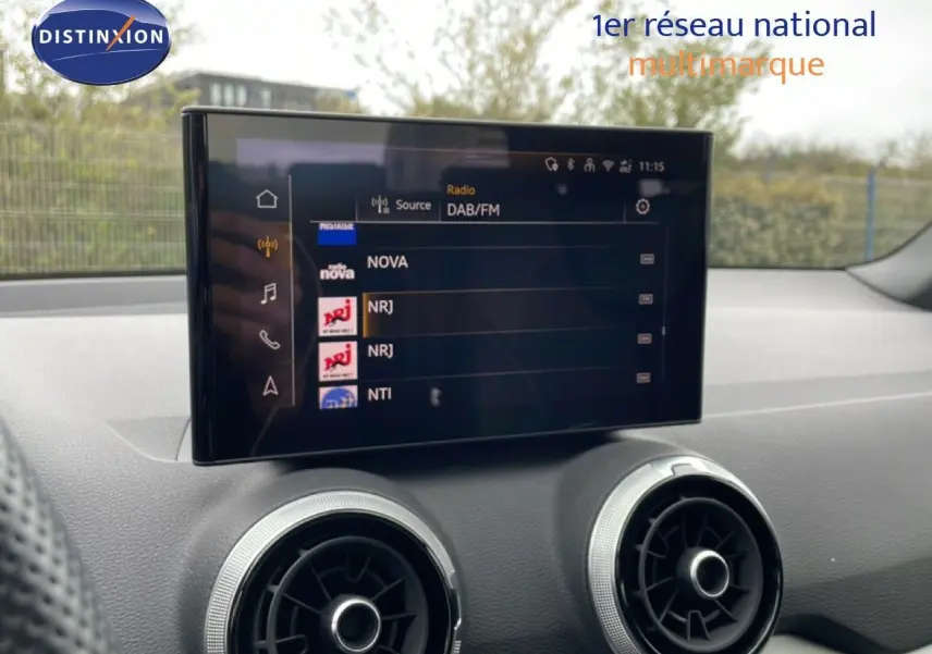 Écran tactile central affichant les stations radio, avec aérateurs ronds argentés sur tableau de bord noir.