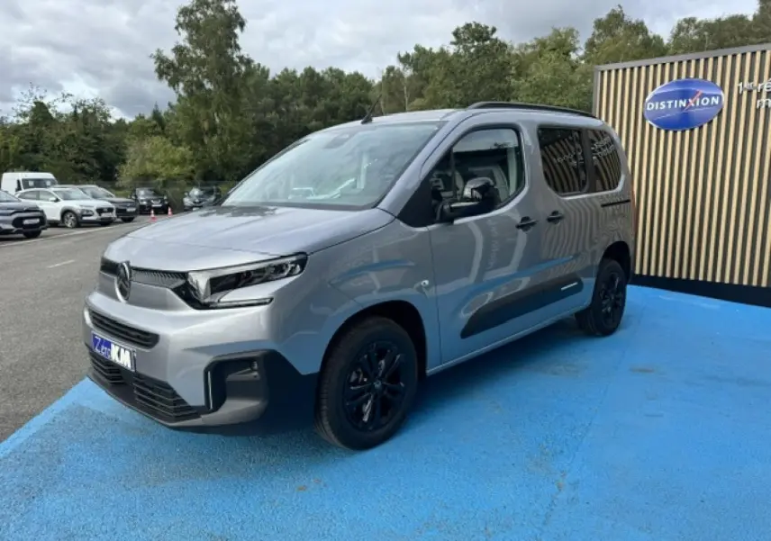 Citroën Berlingo gris acier en 3/4 avant droit, avec jantes noires et barres de toit visibles.