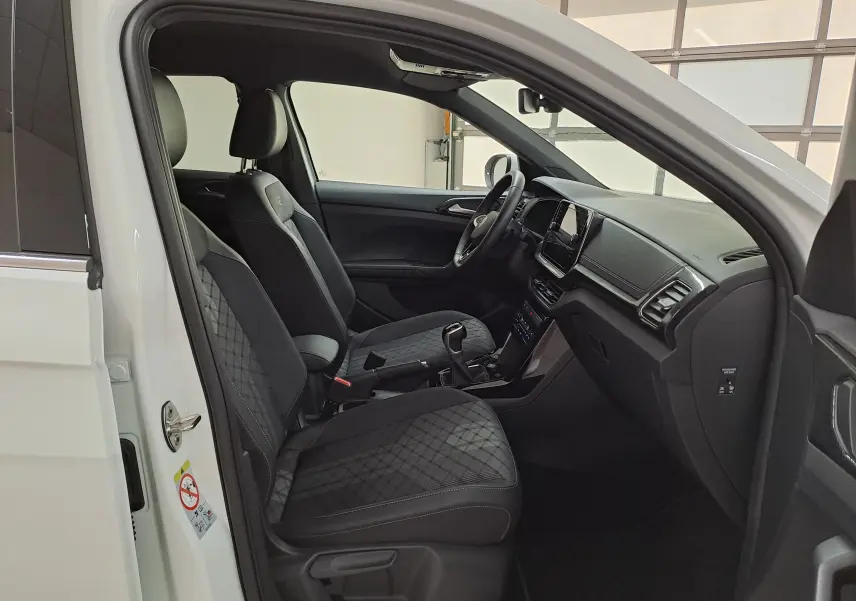 Intérieur du Volkswagen T-Cross blanc 2025 vu côté gauche, avec sièges R-Line en tissu noir à motif losange et tableau de bord moderne.