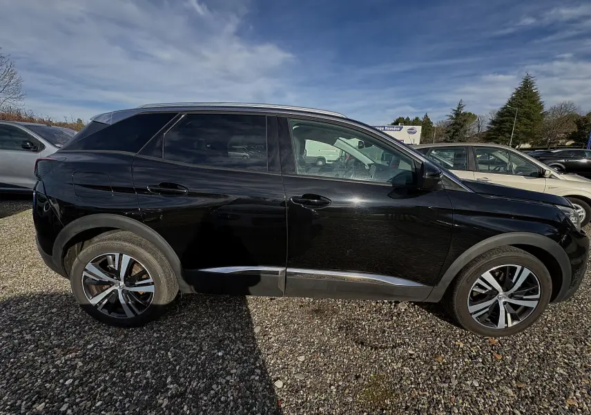 Vue de profil côté gauche d’un Peugeot 3008 noir de 2019 garé sur un sol gravillonné sous un ciel partiellement nuageux.