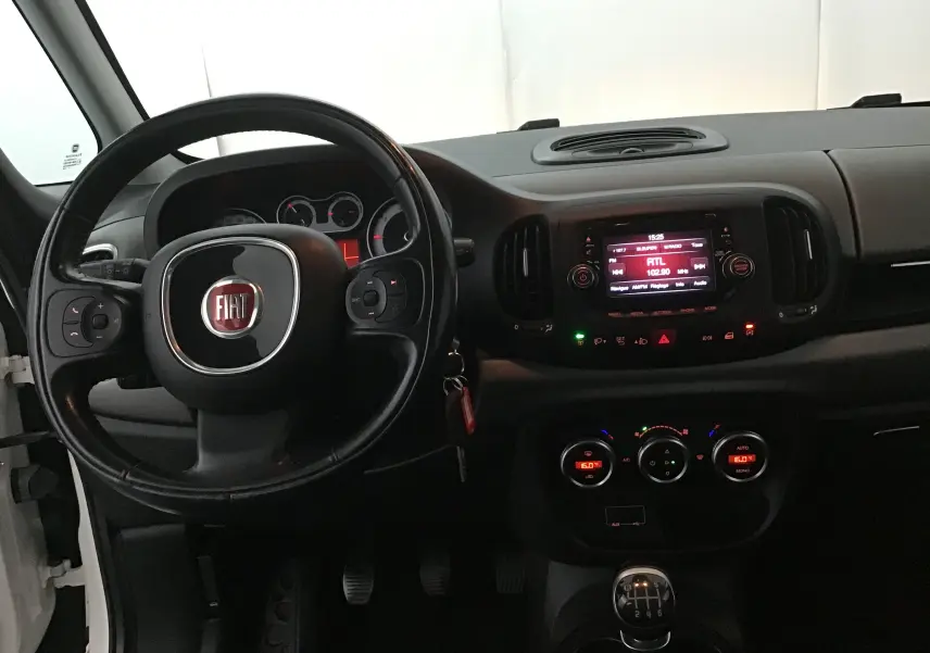 Intérieur de la Fiat 500L blanche vue de face, tableau de bord avec volant, écran multimédia et boîte manuelle visible.