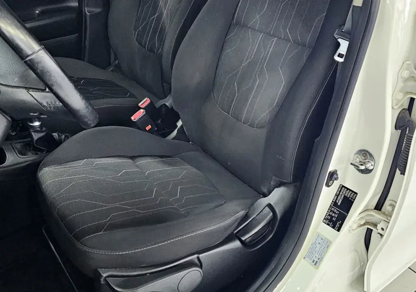 Vue intérieure côté conducteur d'une Kia Picanto II blanche de 2016, sièges noirs avec surpiqûres et volant visible.