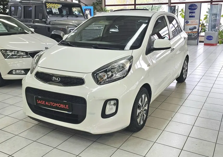 KIA Picanto blanc vue 3/4 avant droit en showroom avec plaque SAGE AUTOMOBILE visible