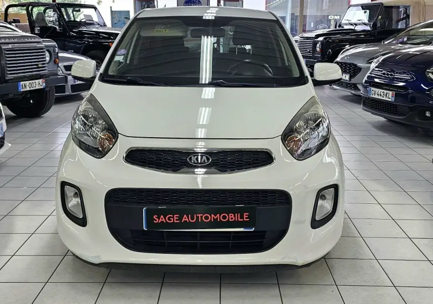 Vue avant d'une KIA PICANTO II blanche de 2016 en showroom, avec calandre noire et phares arrondis visibles.